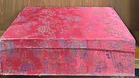Closed decorative red fabric-covered box with floral and traditional Chinese motifs, showing the top view.