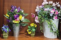 Four artificial floral arrangements displayed on a wooden shelf: metal vase with purple and yellow tulips, white ceramic planter with pink flowers and ivy, two small floral paper wrapped flower arrangements.
