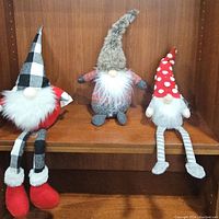 Three plush gnomes displayed on a wooden shelf with different colored hats and outfits.
