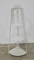Front view of white metal 3-tier corner plant stand showing perforated triangular shelves and decorative scrollwork on top.