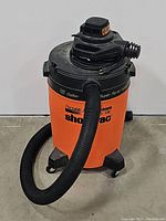 Front view of the orange 16 gallon Shop Vac with black hose attached and mounted on black caster wheels.