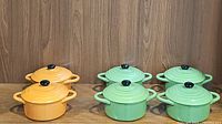 Photo showing 2 orange and 4 green stoneware mini bakers with lids, all with black knob handles, set against a wood background.