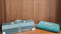 Photo of one vintage metal toolbox showing wear, rust, and tape on corners and one green plastic rivet kit case with latch closure on wooden shelf
