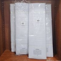 Seven white tulle fabric bolts shrink-wrapped in clear plastic with warning labels, arranged standing upright on a wooden shelf.