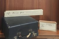 Black carrying case for the typewriter with worn handle and metal latches, with boxed Tavern pipe and Bicentennial brick visible behind.