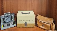 Vintage Kodak Brownie 8 movie camera projector, case, and brown leather camera bag, all arranged on wooden shelf.