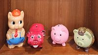 Front view of four piggy banks arranged side by side on a wooden shelf, showing the variety of styles and colors.