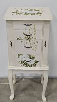 Front view of white hand painted wooden jewelry armoire with floral designs on drawers and top. Shows 4 drawers closed and curved legs.