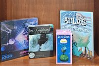 Four items arranged: Star Trek 1000-piece puzzle, Jean Guichard Lighthouses 500-piece puzzle, Young People's Atlas of the United States book, and a hot air balloon musical box in original box.