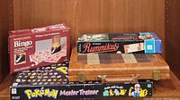 Photo of Rummikub, Bingo, and Pokemon Master Trainer game boxes stacked, showing game titles and box condition.