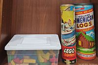 Plastic container with assorted building blocks, LEGO System tube, and vintage Halsam American Logs cylindrical container placed side by side on a wooden shelf