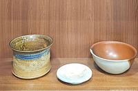 Three ceramic pieces displayed against wooden background: a blue and tan crock, white and brown bowl, and white saucer.