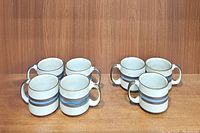 Seven vintage Otagiri Horizon ceramic coffee mugs arranged in two rows on wooden surface, showing the side profile with handles visible. They have beige speckled glaze with brown and blue horizontal stripes.