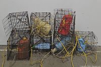Seven used metal square crab pots with multiple colored buoys and attached yellow ropes, arranged on a concrete floor.