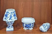 Three blue and white porcelain items positioned on a wooden shelf including a candle holder, a square planter, and a Dutch shoe planter.