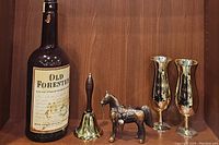 Front view of large Old Forester whisky bottle, vintage brass hand bell with wooden handle, bronze horse figurine, and two gold hurricane glasses on wood shelf.