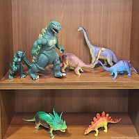 Photo showing seven used plastic dinosaur toys placed on two shelves including Godzilla and various dinosaur species.