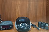 Photo of Sony clock radio, GPX CD radio boombox and Nikon film camera arranged on a wooden surface