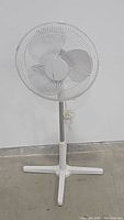 Full view of the white enameled metal standing floor fan with four-legged base against a white wall.