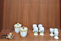 View of all items on a wooden shelf including duck couple egg cups, plain white egg cups, vintage Italian jam jars with lids, and bird-handle spreaders.
