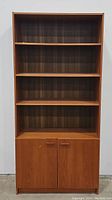 Front view of the tall teak bookcase showing three shelves and closed cabinet doors below.