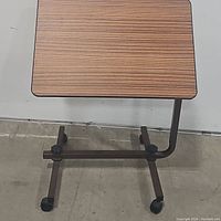 Image showing the adjustable overbed table from the side with visible wood grain tabletop, metal frame, and rolling casters.
