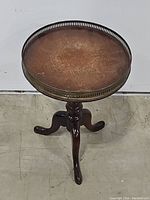 Front view of round wooden table with decorative brass edging and three cabriole legs on turned pedestal base.