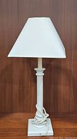 Full view of white candlestick table lamp with shade and coiled power cord on base against dark wood background