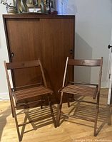 Front view of two wooden folding chairs placed side by side on hardwood floor against cabinetry.