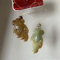 Two jade koi fish carvings displayed side by side on a white surface with a red pouch visible. Each carving has a bead and hanging string attached.