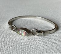 Sterling silver thin bangle bracelet with central eye-shaped opalescent stone, laying flat on fabric background.