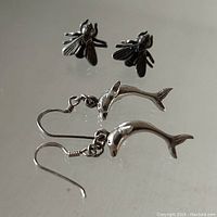 Photo showing a pair of sterling silver fly stud earrings and a pair of sterling silver dolphin dangle earrings on a reflective surface.
