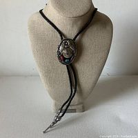 Bolo ties displayed on mannequin neck showing oval decorative slide with eagle and turquoise/coral inlay and braided leather cord with metal tips.