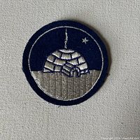 Close-up photo of embroidered WWII Operation Eskimo sleeve patch featuring igloo with smoke plume on dark blue background