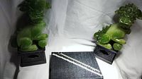 Two bracelets laid out between two green foo dog jewelry display statues, one is a white crystal stone set bracelet, the other a thick chain link bracelet.