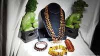 Front view of the five-strand amber necklace displayed on a black bust, accompanied by three amber bracelets in different styles and a black jewelry display base with green foo dog statues on each side.