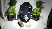 Full lot containing semi-precious stone necklace displayed on black bust with green quartz pendant, Tacori ring on beige ring display, and multicolored polished agate beads bracelet.