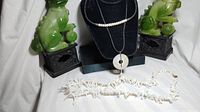 Two Foo Dog jewelry display units flank a black necklace stand holding two necklaces: a pearl necklace on top and a pendant necklace with an abalone shell circle and pearl drop. Below on the surface is a white coral necklace laid flat.