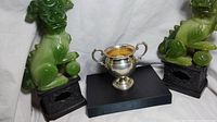 Front and side view of the sterling silver weighted sugar bowl dish placed on a black base, flanked by two green foo dog jewelry display units.