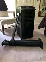 Front and side view of black Danby portable air conditioner with a flexible vent hose and window installation kit part lying next to it on a carpeted floor in a residential room.
