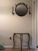 Half round glass table with decorative black metal frame and leaf design, placed under matching round wall mirror and two leaf design wall sconces.