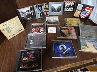 Photo of multiple rock music CDs laid out on a wooden table showing artists such as The Rolling Stones, U2, Creedence Clearwater Revival, John Fogerty, and others