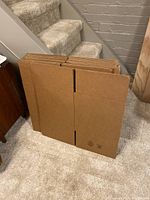 Photo showing flat packed cardboard boxes stacked upright on carpeted floor near stairs.