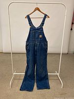 Full front view of Carhartt blue denim overalls hanging on a rack