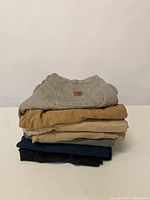 Stack of 7 men's pants in various colors including grey, tan, beige, navy blue, black, and brown, folded on a white surface.