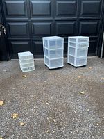 Three white plastic storage units outdoors on pavement: two three-drawer units on castors and one smaller four-drawer tabletop unit.