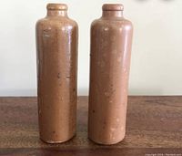 Front view of two small antique stoneware bottles, showing their cylindrical shape and narrow necks with rounded rims, with surface spotting and some wear marks.