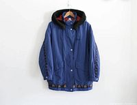 Front view with hood attached, showing blue jacket with black hood, snap button and zip closure, embroidered Fila logo on chest, Aztec pattern trim on sleeves and bottom hem, and two large snap button flap pockets on front.