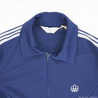 Close-up of collar showing Adidas label and embroidered Montreal Olympics logo on blue fabric with white stripes on sleeves visible.