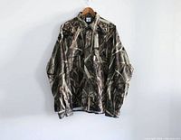 Front view of long-sleeve camouflage hunting shirt hanging on wooden hanger against white wall.